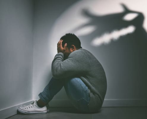 Shot Of A Young Man Sitting In The Corner Of A Dark Room With A Scary Figure On The Wall
